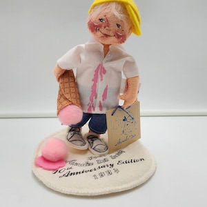 May include: A collectible doll wearing a yellow cap, white shirt with pink stains, and blue shorts, holding a waffle cone with a pink ice cream scoop. The doll stands on a circular base printed with Annalee Doll 10th Anniversary Edition 1993.