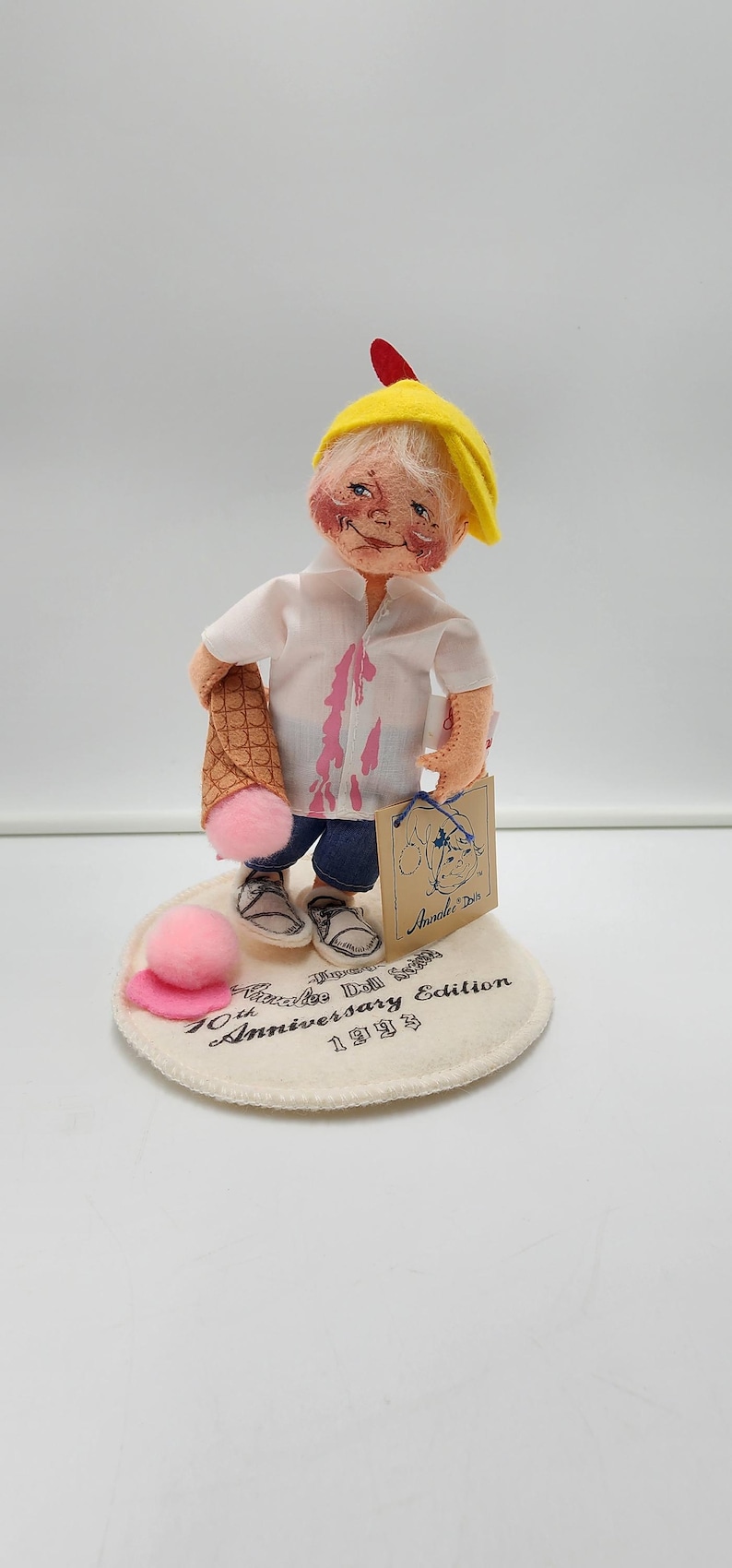 May include: A collectible doll wearing a yellow cap, white shirt with pink stains, and blue shorts, holding a waffle cone with a pink ice cream scoop. The doll stands on a circular base printed with Annalee Doll 10th Anniversary Edition 1993.