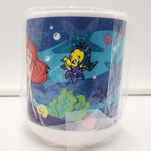May include: A white ceramic mug wrapped in clear plastic packaging featuring a colorful underwater scene with a red-haired character, a yellow and blue fish, green seaweed, and small bubbles against a dark blue background.
