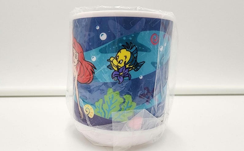 May include: A white ceramic mug wrapped in clear plastic packaging featuring a colorful underwater scene with a red-haired character, a yellow and blue fish, green seaweed, and small bubbles against a dark blue background.