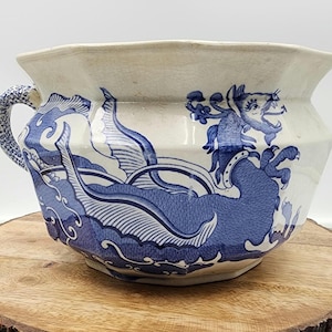 May include: Cream-coloured ceramic bowl featuring a blue dragon and wave pattern. The bowl has a handle and a wide, scalloped rim. The dragon is intricately detailed, and the waves add a dynamic element. The bowl is placed on a wooden surface.