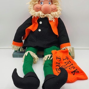Puede incluir: Muñeco de gnomo de Halloween con barba y pelo de paja, vestido con abrigo negro, chaleco naranja, pantalones verdes y zapatos negros. El muñeco sostiene una bolsa naranja con la inscripción "Trick or Treat".
