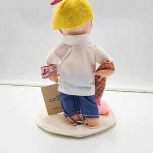 May include: A doll wearing a yellow cap with a red propeller, a white short-sleeved shirt, and blue trousers. The doll holds a brown textured object and a pink item. A tag reads Annalee '93 Made in Meredith, N.H. U.S.A. and another tag is attached.