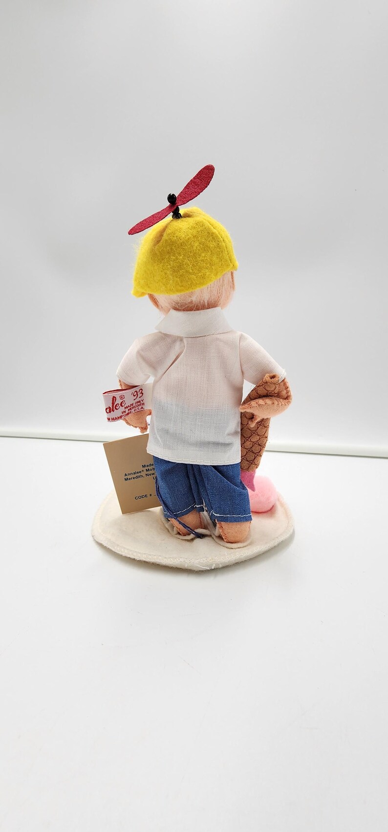May include: A doll wearing a yellow cap with a red propeller, a white short-sleeved shirt, and blue trousers. The doll holds a brown textured object and a pink item. A tag reads Annalee '93 Made in Meredith, N.H. U.S.A. and another tag is attached.