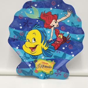 May include: A shell-shaped plate featuring a blue underwater scene with Ariel, Flounder, and Sebastian from Disney's The Little Mermaid. The design includes starfish, bubbles, and a yellow rectangular logo with the text Disney's The Little Mermaid.