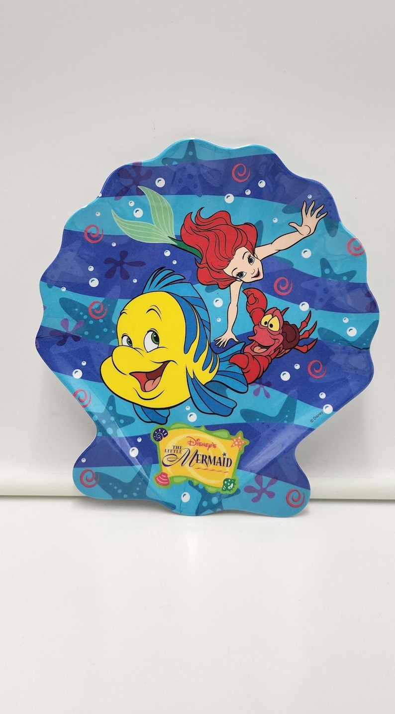 May include: A shell-shaped plate featuring a blue underwater scene with Ariel, Flounder, and Sebastian from Disney's The Little Mermaid. The design includes starfish, bubbles, and a yellow rectangular logo with the text Disney's The Little Mermaid.