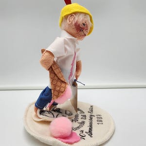 May include: A collectible doll wearing a yellow cap with a red feather, a white collared shirt, and blue trousers. The doll holds a brown waffle cone with a pink ice cream scoop. It stands on a circular base printed with Annalee Doll 10th Anniversary Edition 1993.