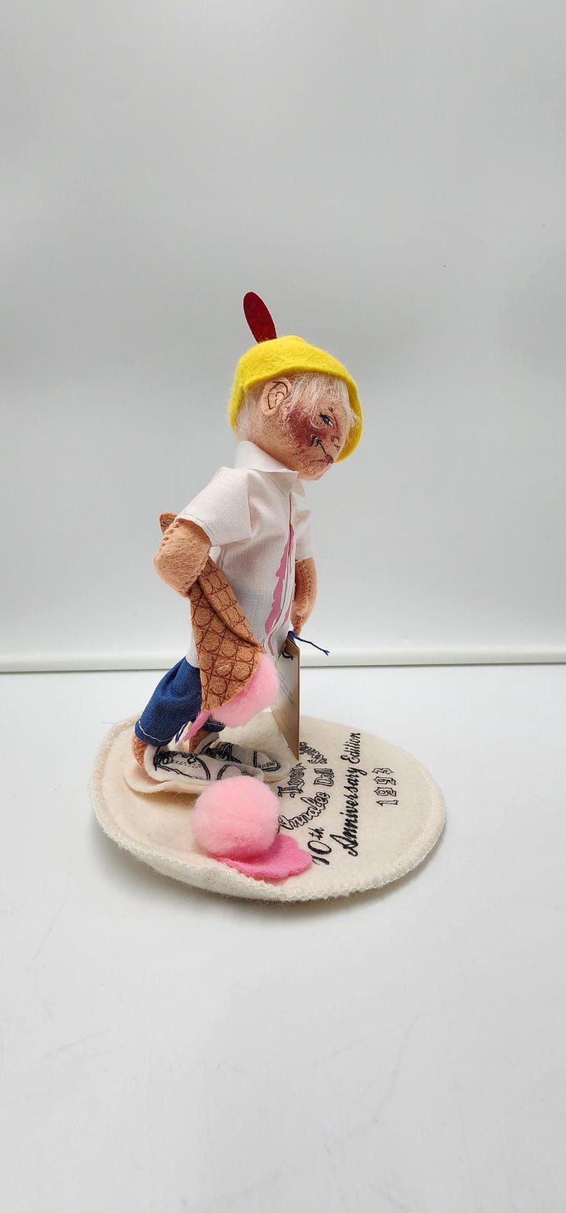 May include: A collectible doll wearing a yellow cap with a red feather, a white collared shirt, and blue trousers. The doll holds a brown waffle cone with a pink ice cream scoop. It stands on a circular base printed with Annalee Doll 10th Anniversary Edition 1993.