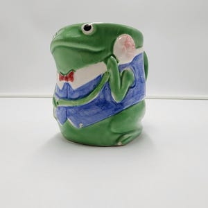 May include: A ceramic mug shaped like a green frog wearing a blue waistcoat over a white shirt with a red bow tie. The frog has large black eyes and a hand raised to its head. The mug features a glossy finish and a sculpted three-dimensional design.