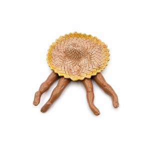 May include: A ceramic sculpture featuring a yellow woven straw hat with a scalloped edge, positioned above four brown, realistic-looking human legs that extend outward from beneath the brim, showcasing detailed knees, ankles, and feet.