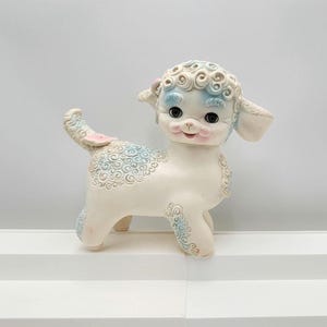 May include: A vintage lamb figurine in off-white with blue and pink accents. The lamb has a curly head, ears, and body details. Its face is pink with black eyes. The lamb is standing on a white surface.