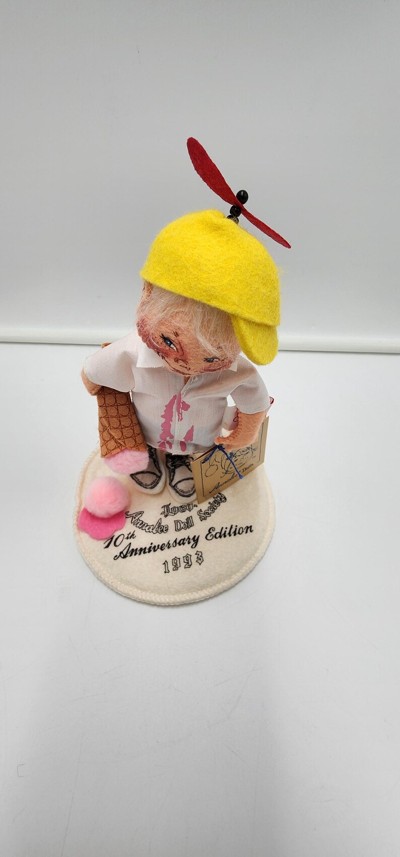 May include: A collectible doll wearing a yellow propeller cap, white shirt with pink accents, and holding an ice cream cone. The doll stands on a circular base printed with Annalee Doll Society 10th Anniversary Edition 1993. A pink pom-pom sits on the base.