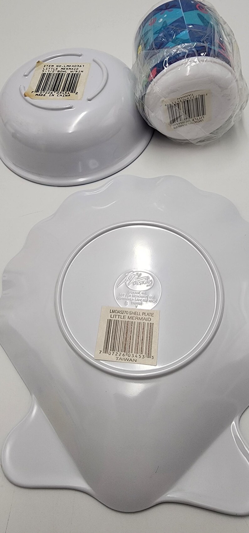 May include: A white plastic shell-shaped plate, a white bowl labeled 5-1/2 inch bowl with rim, and a plastic-wrapped tumbler featuring Little Mermaid graphics. Each item has a barcode sticker and manufacturer markings on the underside.