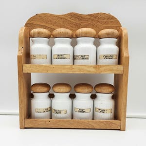 May include: A wooden spice rack with eight white ceramic jars, each topped with a wooden lid. The jars are labeled with various spice names. The rack has two shelves and a decorative top.