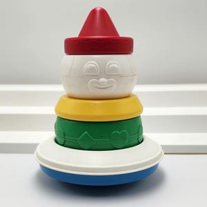 Könnte beinhalten: Ein stapelbares Spielzeug bestehend aus einem roten kegelförmigen Oberteil, einem weißen Kopfteil mit geformtem lächelndem Gesicht, einem gelben Ring, einem grünen Ring mit geprägten geometrischen Formen und einer breiten weißen Basis auf einem blauen Boden.