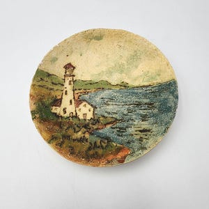 May include: A round ceramic dish featuring a hand-painted coastal scene. The design depicts a white lighthouse standing on a grassy cliff overlooking a textured blue ocean. The color palette includes shades of green, brown, and blue on a cream base.