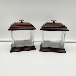 May include: Two clear glass jars with dark brown wooden lids and bases. Each jar has a square shape with a stepped lid and a small gold-coloured knob on top. The jars are empty and sit on a white surface.