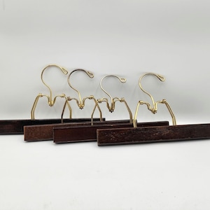 May include: Four wooden skirt hangers with gold-colored metal hooks and clips. The hangers have a dark brown finish and are arranged in a row against a white background. The clips are designed to hold skirts or pants.