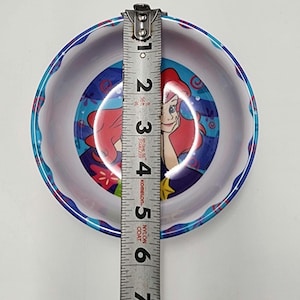 May include: A round bowl featuring a graphic of a red-haired character with a blue background. A metal measuring tape is placed across the center, showing the bowl has a diameter of approximately 5.5 inches. The rim has a blue and white pattern.