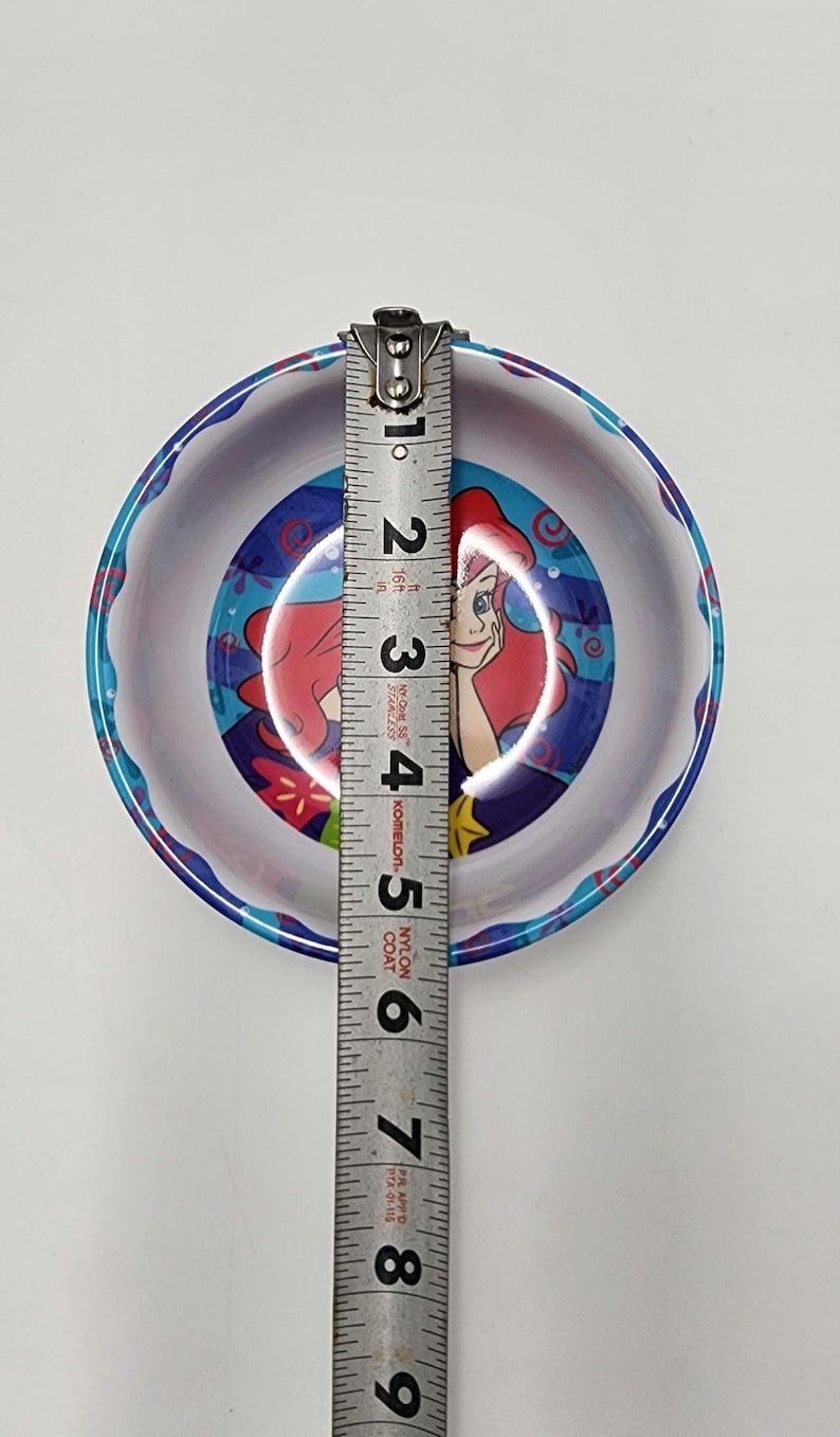 May include: A round bowl featuring a graphic of a red-haired character with a blue background. A metal measuring tape is placed across the center, showing the bowl has a diameter of approximately 5.5 inches. The rim has a blue and white pattern.
