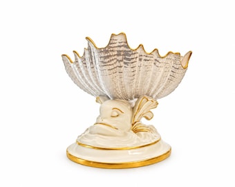 May include: A ceramic bowl shaped like a scalloped shell with a silver-toned interior, supported by a cream-colored base featuring a sculpted fish figure. The rim of the bowl and the circular base are accented with a thin, polished gold-colored trim.