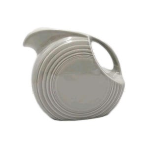 Puede incluir: Una jarra de cerámica de color gris claro con un cuerpo redondeado en forma de disco que presenta anillos concéntricos en relieve que irradian desde el centro. La jarra tiene un asa curva en el lado derecho y un pico vertedor ancho y cónico.