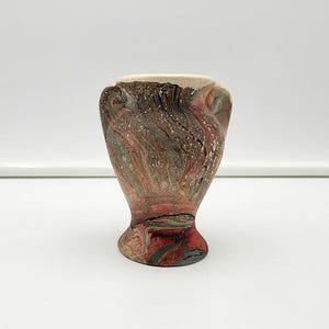 May include: A small, decorative vase with a marbled design in shades of red, brown, and green. The vase has a wide, rounded body with two handles and a cream-coloured rim. The base is a darker shade of red.