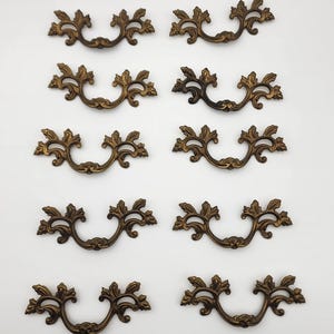 May include: A set of ten matching antique brass-toned metal drawer pulls arranged in two vertical columns. Each pull features a curved, ornate design with symmetrical leaf-like flourishes extending from the center mounting points.