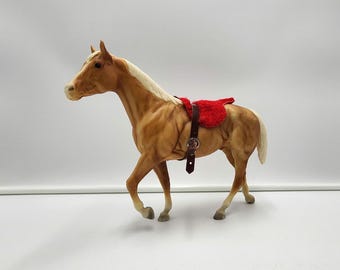 以下が含まれることがあります： 明るいクリーム色のたてがみと尾を持つパロミノ種の馬のフィギュア。馬には銀色のバックルが付いたダークブラウンのレザー製サドルストラップが装着されており、背中には鮮やかな赤色の質感のあるサドルパッドが乗せられています。