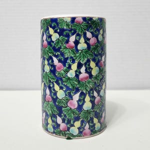 Puede incluir: Un jarrón de cerámica cilíndrico con fondo azul y un patrón repetido de hojas verdes y calabazas amarillas, rosas y blancas.