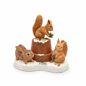 May include: A decorative figurine featuring two brown squirrels and a brown rabbit on a white, snow-covered base. One squirrel sits atop a brown terracotta-style pot with green holly leaves and red berries, while the other holds a pinecone.