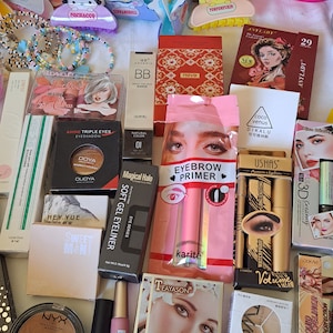 May include: A collection of makeup products, including eyeshadow palettes, eyeliner, mascara, eyebrow primer, and foundation. The products are arranged on a white surface and feature various colors and designs.