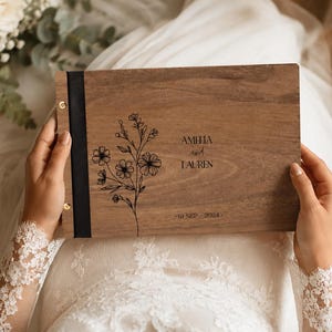 May include: A rectangular wooden photo album with a dark brown wood grain finish. The cover features a black vertical fabric spine with gold metal binding screws. A black floral line art illustration is printed on the left, with the text AMELIA and LAUREN and 10 SEP 2024.