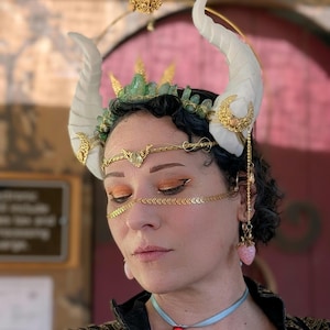 Puede incluir: Un tocado de fantasía con cuernos blancos, una corona dorada con cristales verdes y detalles dorados. El tocado incluye una cadena dorada para la frente y pendientes colgantes con dijes de fresa rosa. Un collar de cinta azul con un colgante de corazón rojo completa el look.