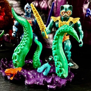 May include: A purple base with two large green tentacles, a small blue tentacle, and an orange frog figure. Behind the base stands a teal action figure with a cat-like face, gold armor, and a yellow sword, alongside a blue-skinned action figure.