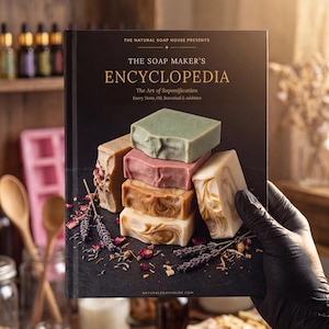 Puede incluir: Un libro de tapa dura titulado The Soap Maker's Encyclopedia: The Art of Saponification. La portada negra muestra una pila de cuatro pastillas de jabón rectangulares en tonos verde, rosa y bronceado jaspeado, rodeadas de lavanda y pétalos de rosa.