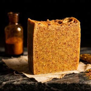 Puede incluir: Una pastilla de jabón rectangular de color marrón dorado con una textura granulada y pequeñas partículas vegetales visibles. El jabón tiene un borde superior irregular y se apoya sobre un trozo de tela clara, junto a un frasco de vidrio ámbar.