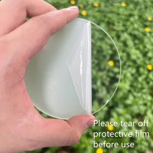 May include: A round, light green object with a clear protective film being peeled off. The text "Please tear off protective film before use" is visible in the lower right corner. The background is a blurred green with yellow flowers.