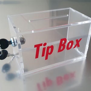 Può includere: Una scatola per mance in acrilico trasparente con una fessura sulla parte superiore e la scritta "Tip Box" in rosso. È dotata di serratura e due chiavi. Progettata per raccogliere mance.
