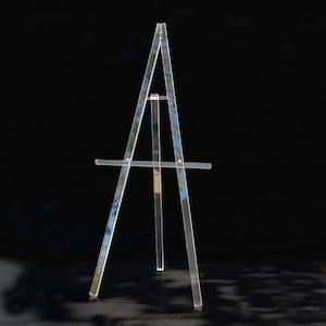 May include: A clear acrylic easel with a three-legged design. The easel has a horizontal support bar and a vertical support beam. The easel is designed to hold a canvas or artwork. The background is black.