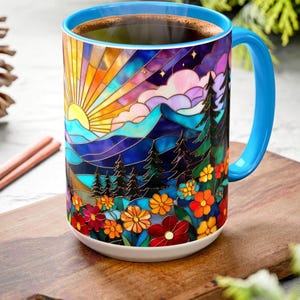 May include: A ceramic coffee mug with a colorful stained-glass design featuring a sunrise over mountains, trees, and flowers. The mug has a blue handle and rim, and is filled with dark coffee. The mug is on a wooden cutting board.