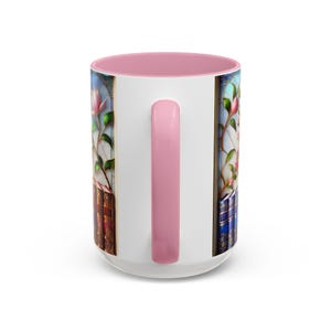 May include: White ceramic mug with a pink interior and handle. The mug features a stained-glass design with pink flowers and books. The handle is a solid pink color. The mug is a standard size for coffee or tea.