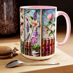 May include: White ceramic mug with a pink handle and rim, featuring a stained-glass design of flowers and books. The floral design includes pink, purple, and white flowers with green leaves. The base of the mug has a row of books. A silver spoon and a macaron are in the foreground.