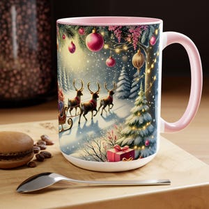 Pode incluir: Uma caneca grande de cerâmica branca com interior e alça rosa. A caneca apresenta uma cena de inverno com renas puxando um trenó, enfeites de Natal e uma árvore de Natal decorada. Uma colher e um macaron estão em primeiro plano.