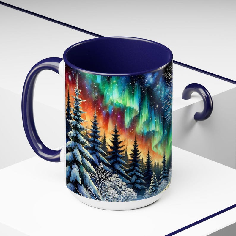 Peut inclure: Mug en c&eacute;ramique blanche avec une anse et un int&eacute;rieur bleu marine. L'illustration vibrante repr&eacute;sente les aurores bor&eacute;ales au-dessus d'une for&ecirc;t enneig&eacute;e. L'&oelig;uvre d'art pr&eacute;sente des nuances de vert, d'orange et de bleu.