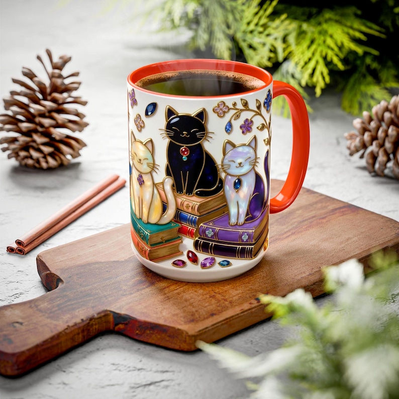 May include: A ceramic coffee mug with a red handle and rim, featuring three stylized cats sitting on books. The cats are black, yellow, and white, adorned with jewels. The mug is filled with coffee and sits on a wooden cutting board.