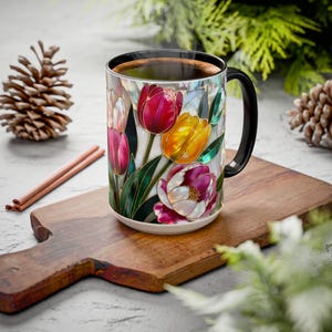 Pu&ograve; includere: Una tazza da caff&egrave; in ceramica con manico e bordo neri, riempita con un liquido scuro. La tazza presenta un design a vetrata di tulipani colorati nelle tonalit&agrave; del rosso, giallo e viola. La tazza &egrave; appoggiata su un tagliere di legno.