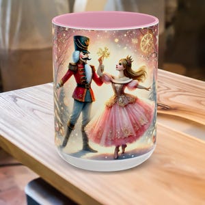 May include: A white ceramic mug with a pink interior features a festive illustration of a Nutcracker and a ballerina. The Nutcracker wears a red jacket and black hat. The ballerina is in a pink dress. The mug is on a wooden surface.