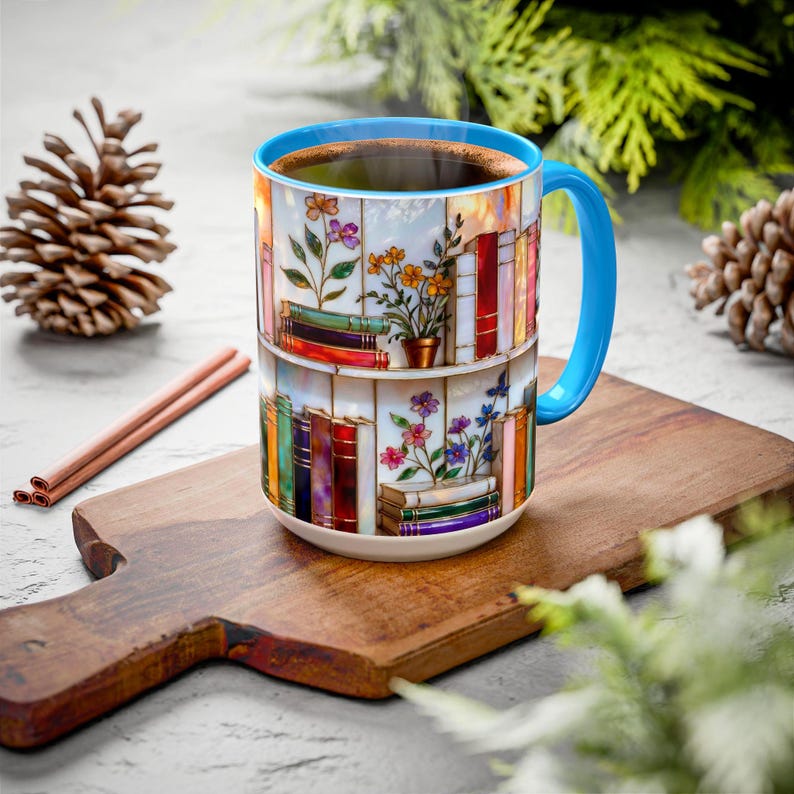 May include: A ceramic coffee mug with a blue handle and rim, filled with dark liquid. The mug features a colourful design of bookshelves with flowers. The mug sits on a wooden chopping board, with a pine cone and cinnamon sticks nearby.