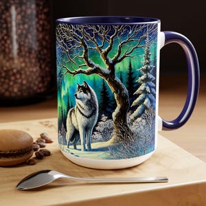 Peut inclure: Mug en c&eacute;ramique blanche avec int&eacute;rieur et anse bleu fonc&eacute;. L'illustration d&eacute;taill&eacute;e repr&eacute;sente un loup dans une for&ecirc;t enneig&eacute;e sous une aurore bor&eacute;ale vibrante. Le motif comprend un grand arbre et des conif&egrave;res.